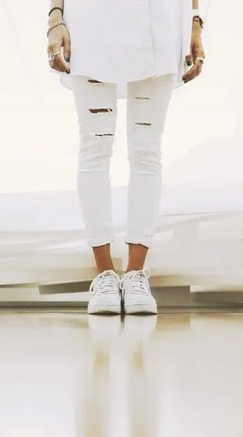 A close-up shot of a pair of clean white minimalist sneakers paired with tailored trousers, showcasing how simple footwear can elevate a casual-chic look. The background is a clean, modern floor.