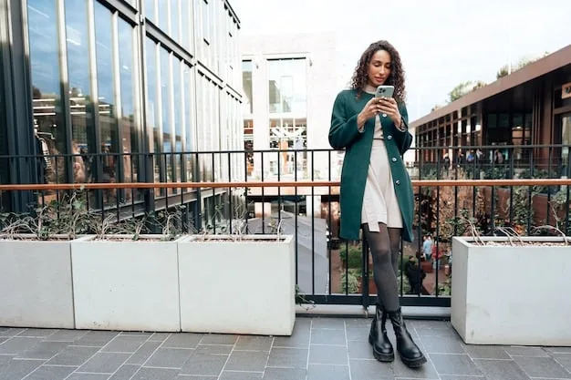 A woman in a long, flowing grey tunic sweater over black leggings, paired with sleek black ankle boots and a minimalist necklace, walking purposefully in a modern city setting at an outdoor cafe.