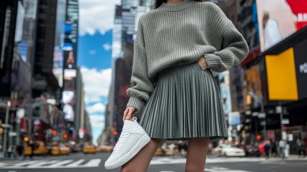 A woman wearing a long, oversized knit cardigan over a floral midi skirt, styled with simple white sneakers. She is casually walking in an urban setting during a mild, overcast day.