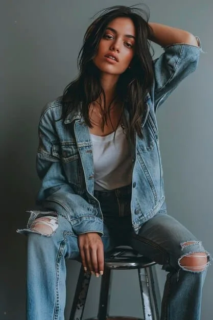 A detailed shot of a relaxed luxe denim on denim outfit, featuring an oversized light-wash denim jacket draped over a cream-colored chunky knit sweater, paired with wide-leg distressed jeans and chunky white sneakers.