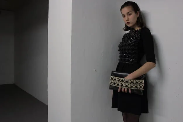 A woman wearing a sleek, minimalist black midi dress with pointed-toe metallic flats and a small, structured clutch bag, standing in a contemporary art gallery.