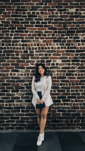 A woman casually leaning against a brick wall, showcasing a sporty chic outfit that combines tailored grey joggers with a black fitted turtleneck, a chic camel trench coat, and white minimalist sneakers. She carries a small, structured crossbody bag.