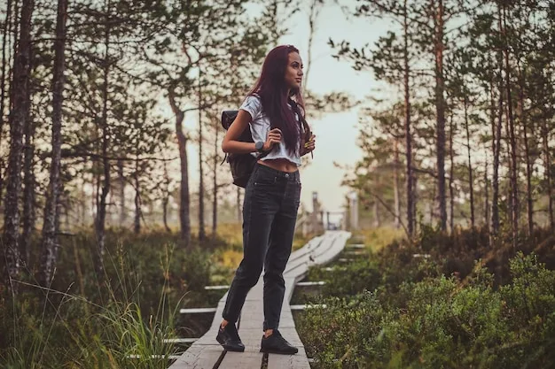 A person standing on a scenic outdoor trail, wearing a layered outdoor enthusiast's refined look: dark olive green technical pants, a cream-colored long-sleeve top, a lightweight charcoal grey puffer vest, and stylish yet practical hiking boots. A durable, minimalist backpack is slung over one shoulder.