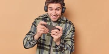 Addicted man plays video games holds smartphone horizontally tries to pass difficult level wears stereo headphones on ears dressed in checkered shirt isolated over beige background. Technology
