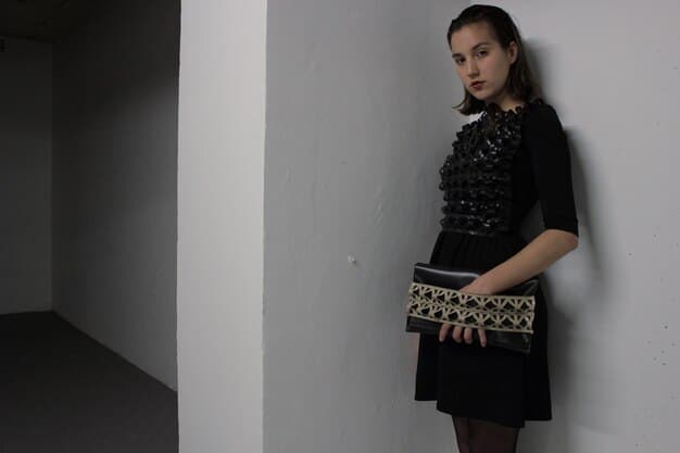 A woman wearing a sleek, minimalist black midi dress with pointed-toe metallic flats and a small, structured clutch bag, standing in a contemporary art gallery.