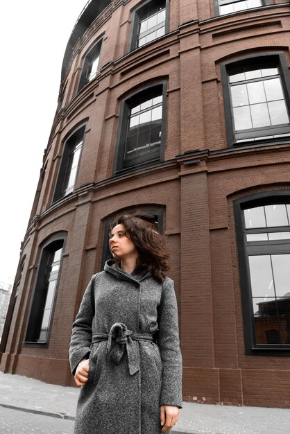 A stylish woman standing in front of a brick building, wearing a camel-colored long coat over a cream-colored turtleneck. She sports a deep maroon pleated skirt and carries a vibrant orange leather tote bag, creating a striking color-blocked effect.