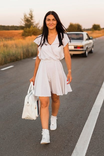 A young woman wearing a flowy white embroidered blouse with bell sleeves, high-waisted flared denim jeans, a wide-brimmed hat, and carrying a fringed cross-body bag, walking on a cobblestone street.